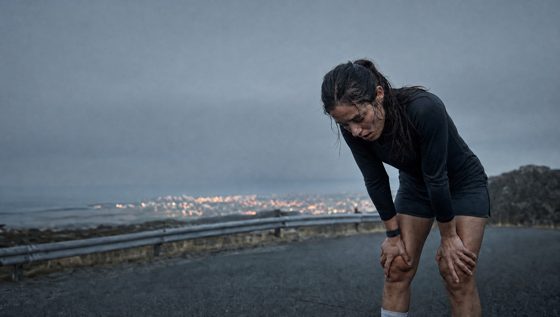 A runner folded at a dusk hilltop crest, chest heaving in clear cold air, city lights far below